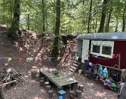 Bauwagen im Wald, davor ein großer Tisch. Darüber hängt eine bunte Girlande.