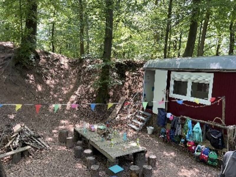 Bauwagen im Wald, davor ein großer Tisch. Darüber hängt eine bunte Girlande.