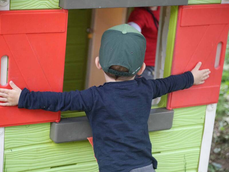 Ein Kind von hinten welches die Arme ganz ausgebreitet hat und vor einem Spielhaus steht und die Fenster schließen möchte